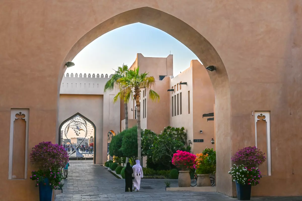 Two people in traditional dress walking through an archway surrounded by flowers and historic architecture in Katara